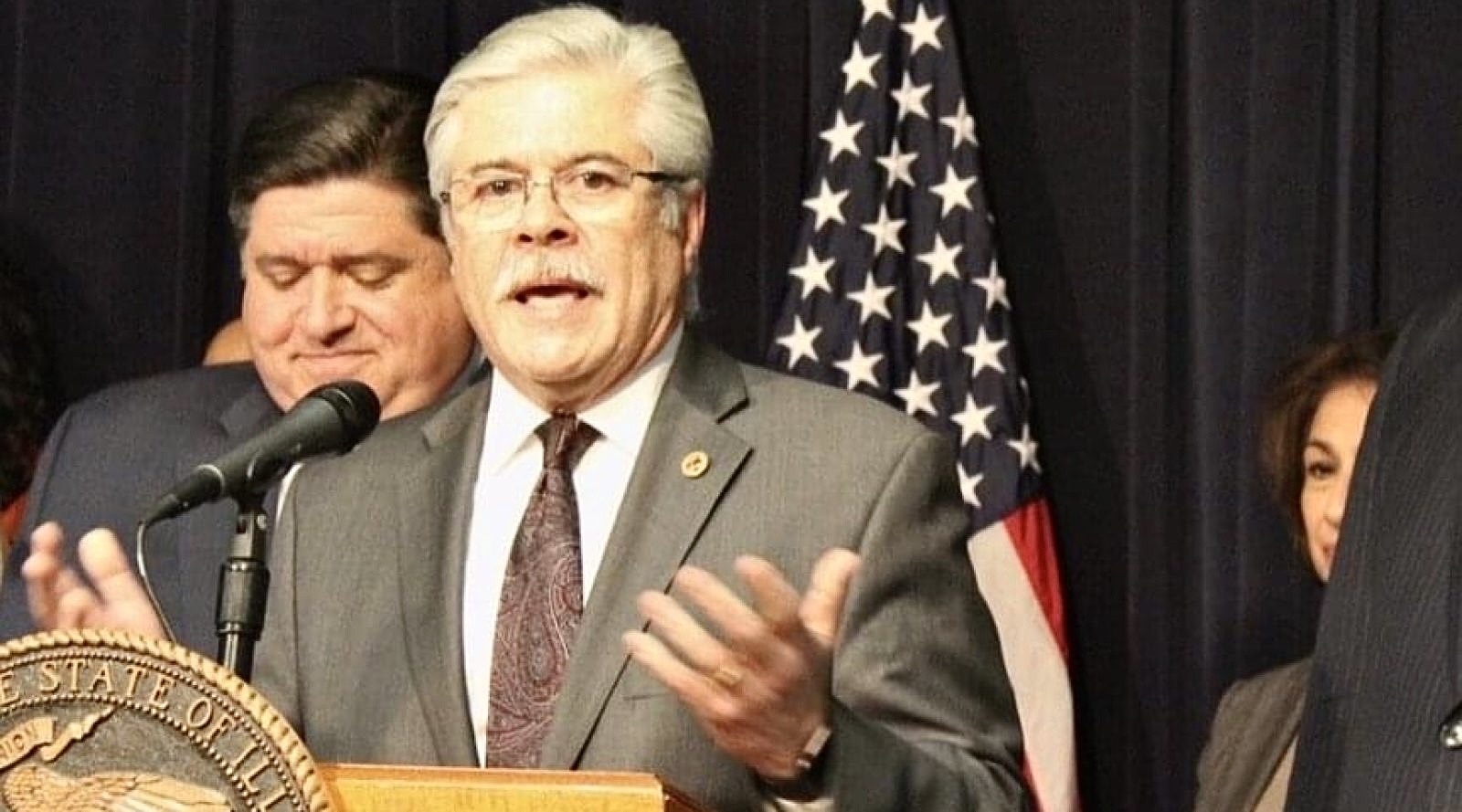 State Representative Fred Crespo speaking at a podium bearing the Illinois state seal.